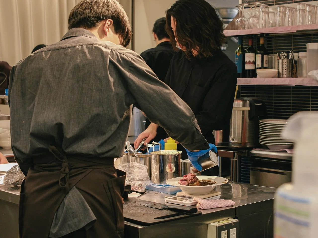 Two restaurant colleagues in dark aprons preparing service together at a stainless-steel kitchen pass.